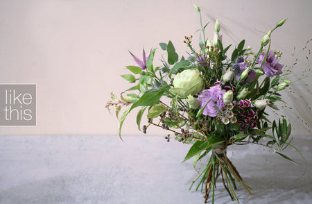 A medium-stemmed lilac and white bouquet with green leaves, arranged in a vase against a neutral background.